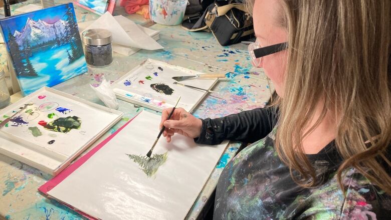 A woman surrounded by art and paints with a mountain forest lake landscape in front of her sits at a desk painting a tree.