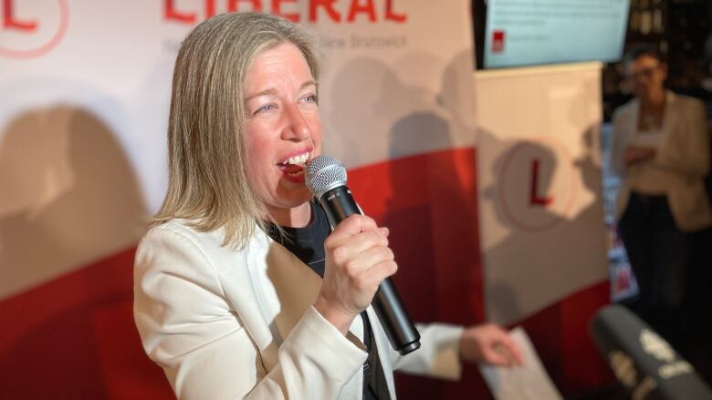 A woman wearing a white blazer speaking into a handheld microphone in front of a red and white banner.