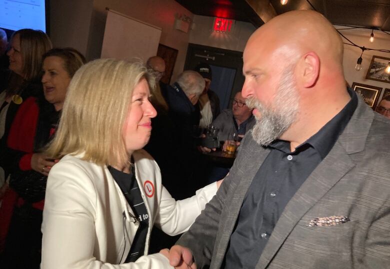 A woman, left, shaking hands with a man, right, with people mingling in the background.