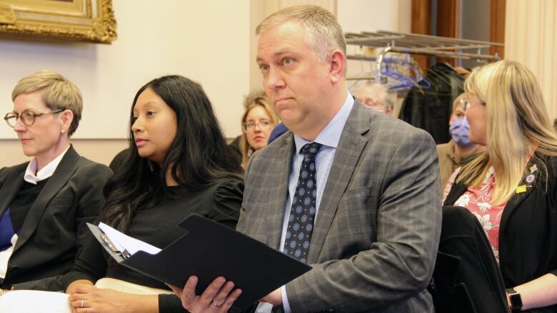 A man sits in a chair, surrounding by others, opening up a folder with his speech inside.