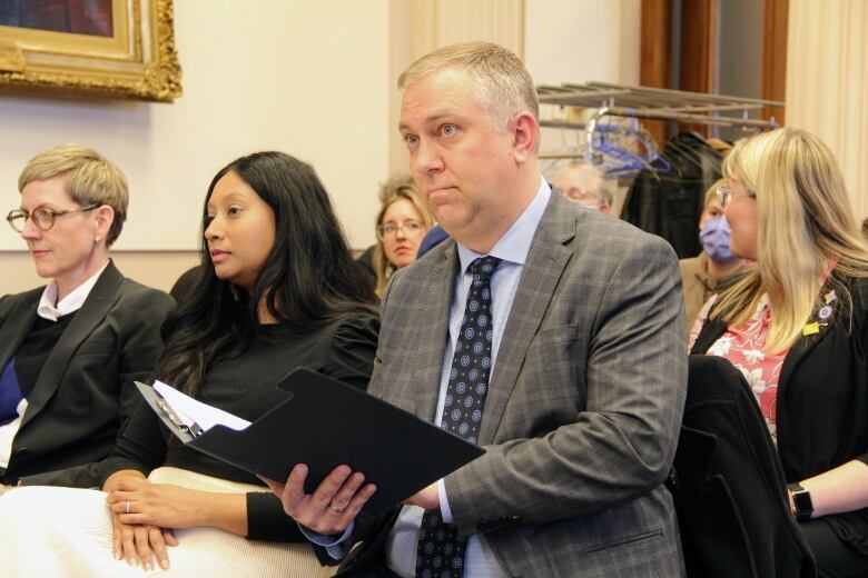 A man sits in a chair, surrounding by others, opening up a folder with his speech inside.