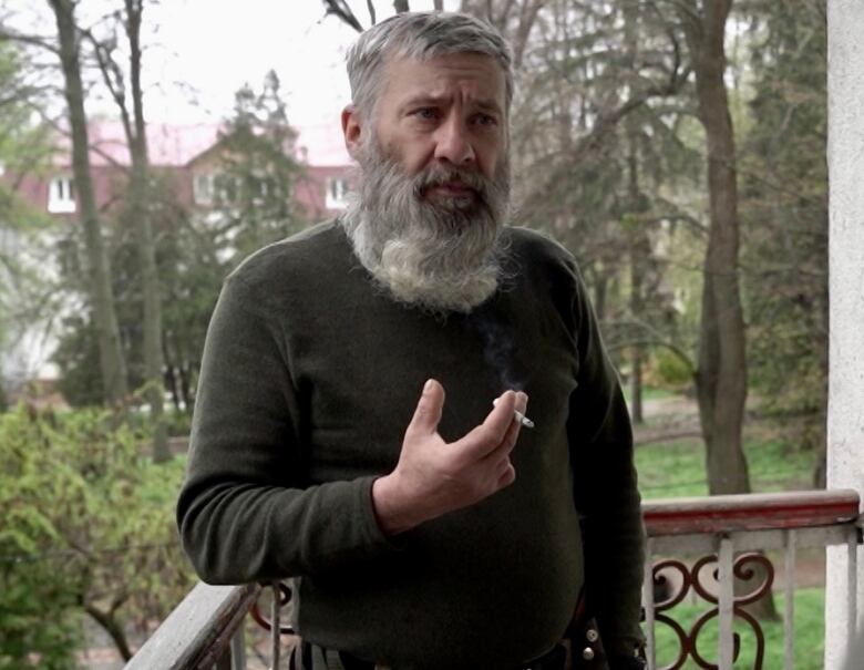 A man in an olive green sweater with short grey hair and a long white beard stands on a balcony holding a cigarette in one hand that is missing two fingers. 