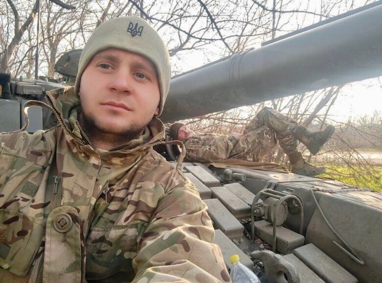 A young soldier in tan fatigues takes a selfie on a tank while another soldier lays on the tank in the background. 