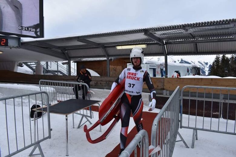 A man in a white and red luge bodysuit carries a red sled away from a luge track on a snowy day.