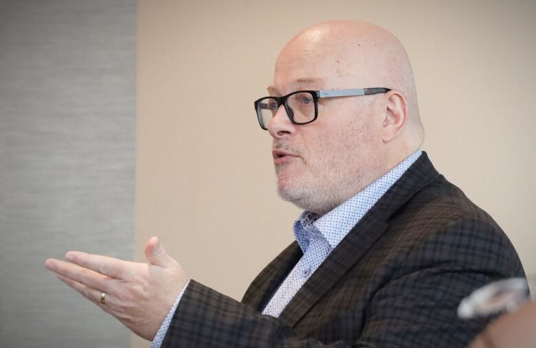 A man with glasses in a brown suit gestures with his left hand while speaking.