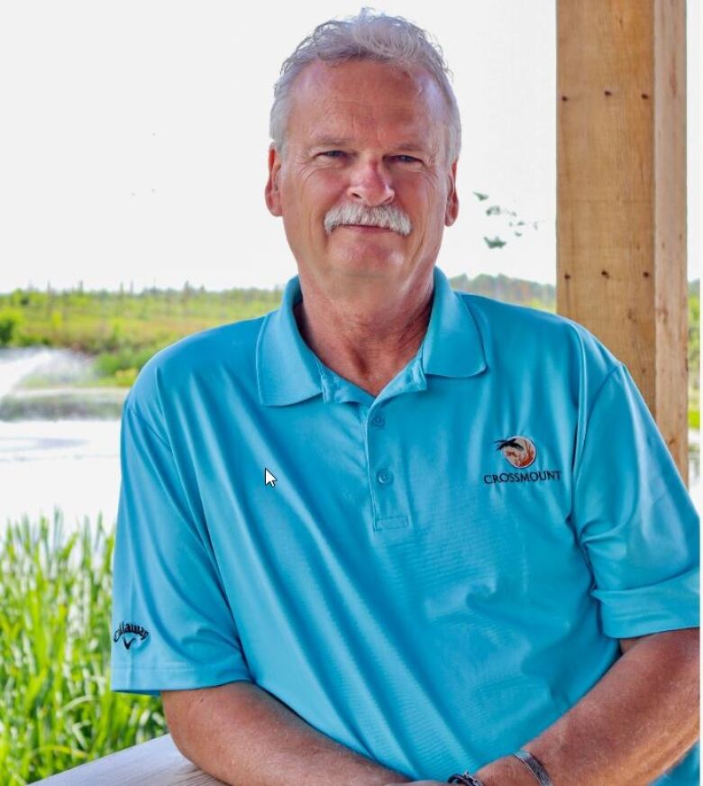 A middle-aged white man with a moustache smiles at the camera. He is standing outside with a lake in the background.