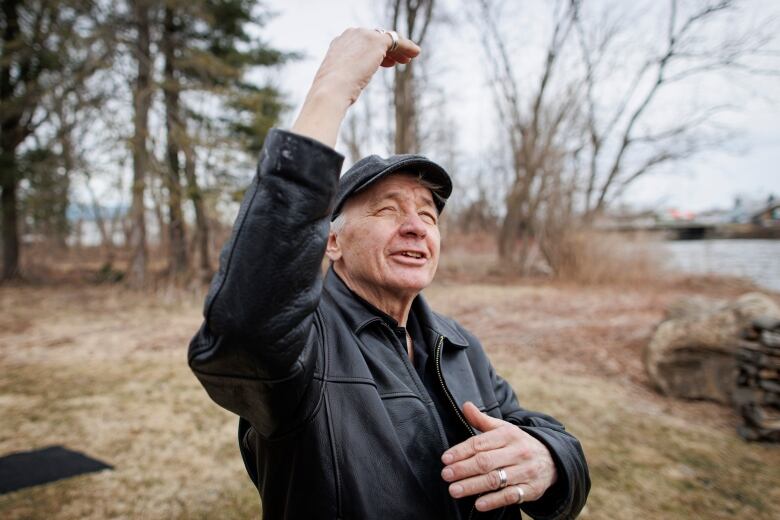 A man wearing a leather jacket and a grey farmers cap gestures with his hands standing on the banks of the St. Lawrence River. 