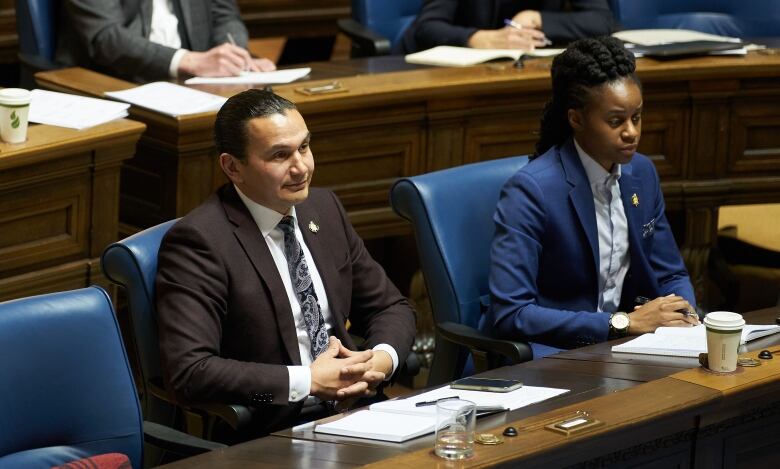 Two people in suits, foreground, sit in front of others in a legislative chamber.