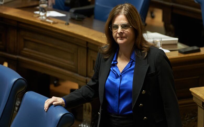 A woman in a black blazer and blue blouse stands in a legislative chamber.