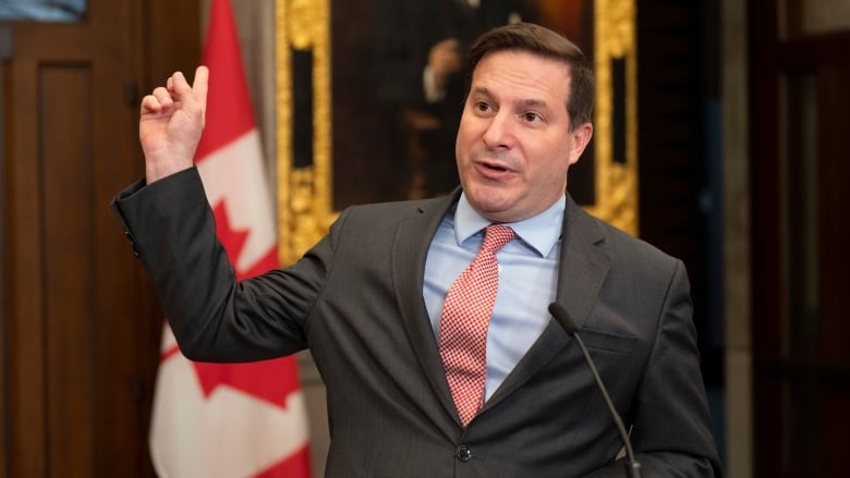 A man in a suit gestures in a House of Commons hallway at a podium.