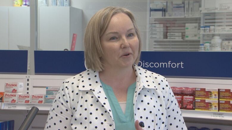 A woman with short blonde hair wears a white jacket with black dots with a green shirt underneath. She is standing in front of a shelf of Tylenol.