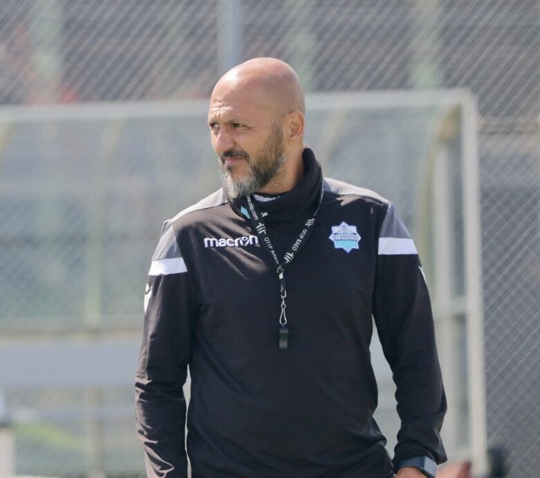 A soccer coach, wearing a team jacket and a whistle around his neck, is photographed.