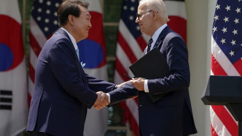 Two men in suits shake hands with the US and Korean flags behind them