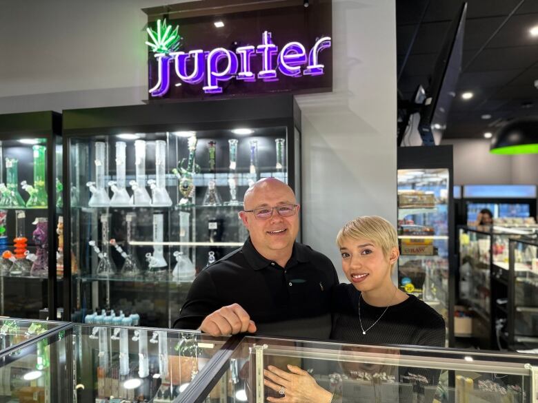 A husband and wife stand in their store surrounded by cannabis glassware. A neon sign that says 