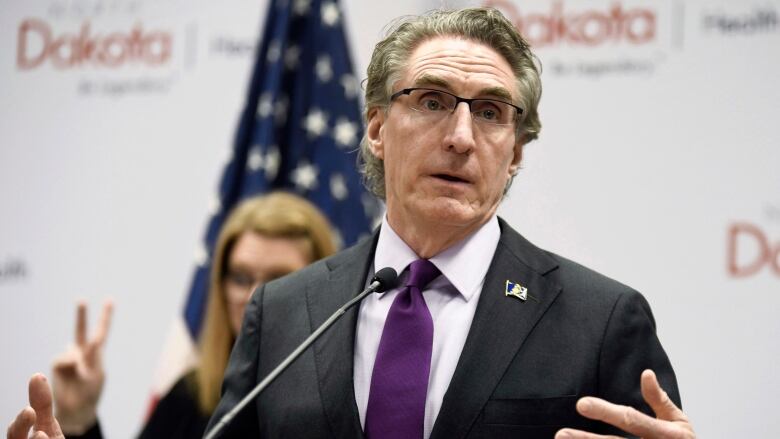 Man in dark suit and purple tie speaks at podium