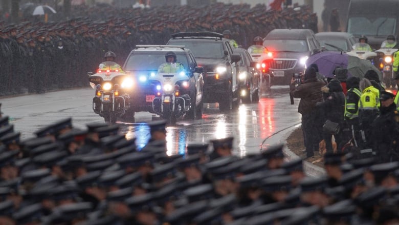 Vehicles drive down a road surrounded by a crow of officers.