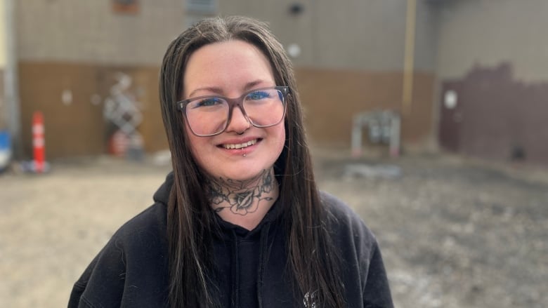 A person stands in a dirt lot, wearing glasses, long dark hear, and a floral neck tattoo.