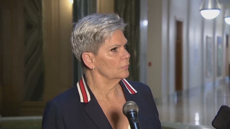 An older woman with short grey hair and pierced ears is wearing a navy blue blazer, the collar of which has a red-and-white striped pattern. She is standing in a rotunda.