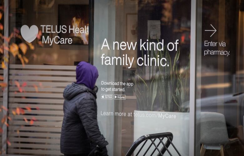 A person passes a health clinic with the words 'Telus Health MyCare' and 'A new kind of family clinic' written on the windows.