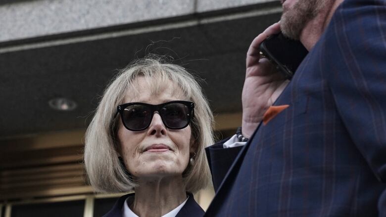 Closeup of a woman wearing sunglasses and standing near a man talking on a cellphone outside a set of doors.