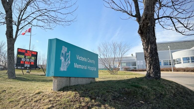 A blue sign on the lawn between two trees indicates Victoria County Memorial Hospital, which is shown in the background, along with a black sign with neon-coloured letters for the urgent treatment centre.