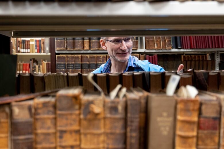 A man pulls out a book on the other side of a bookshelf. The books closest to the camera are yellowed and worn. In the background are more old books.