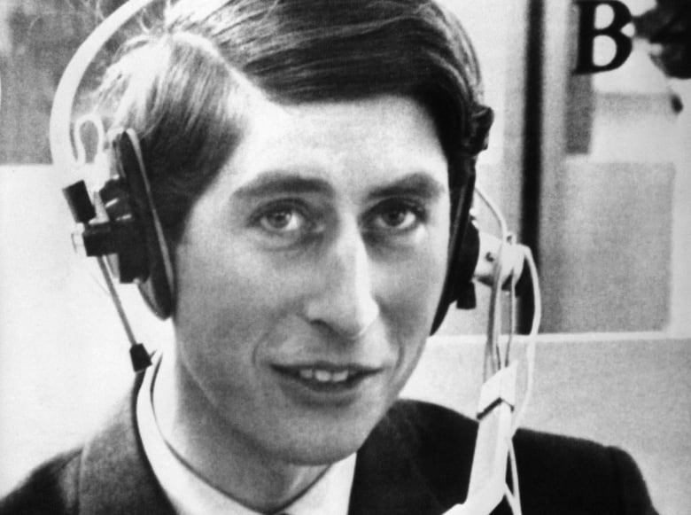 A black and white photo shows a young Prince Charles wearing a headset with a microphone attached. 