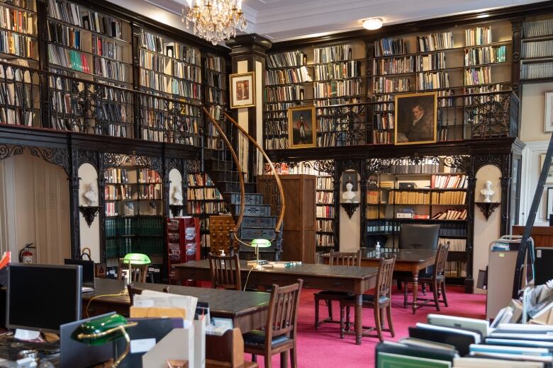 A picture of a two-storey library with a curved staircase, three tables, and rows of books on a pink carpet. There are white busts between book cases.