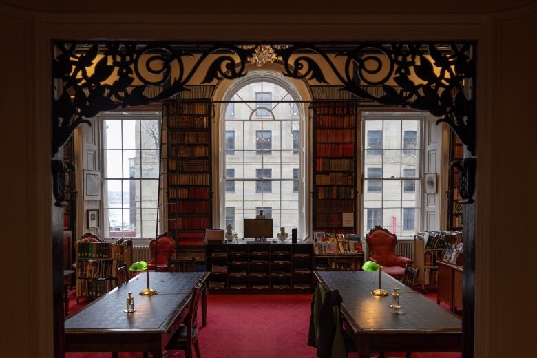 A doorway with wooden details leading to two tables lit by lamps. Behind them are two long bookshelves on the wall, next to three large windows. 