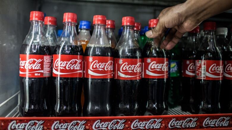 The hand of a man is shown reaching out to grab a bottle of coke from a display