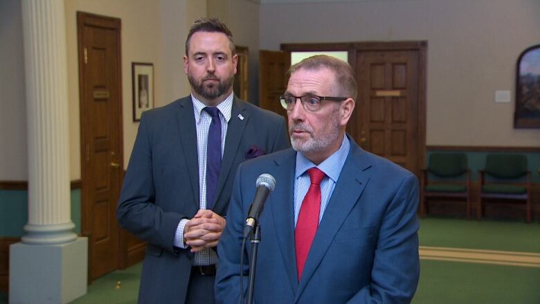 Two men wearing suits stand at a microphone.