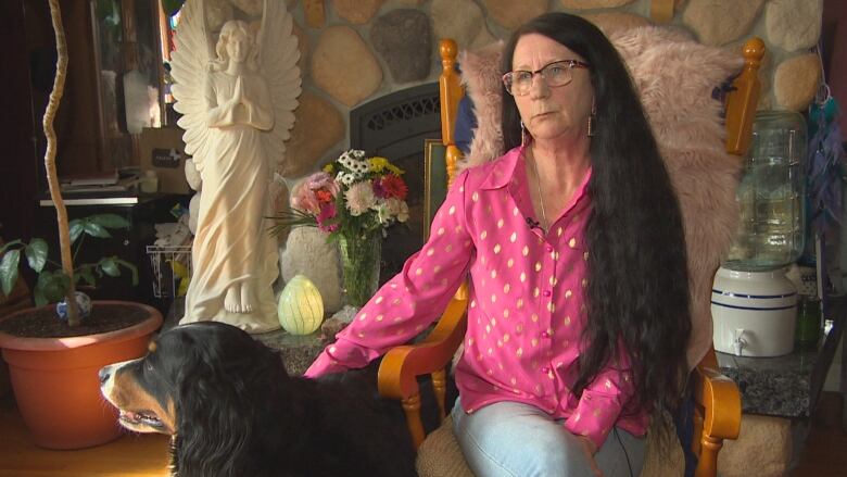 A woman with long dark hair wearing a pink shirt with white polka dots and jeans is sitting on a chair in her living room with her dog beside her. 