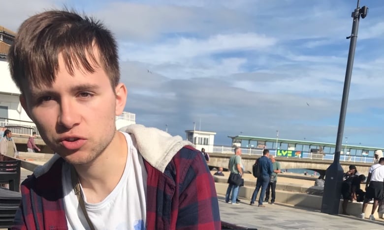 A young man with short brown hair wearing a plaid hoodie, sits at a picnic table under partly cloudy skies. 