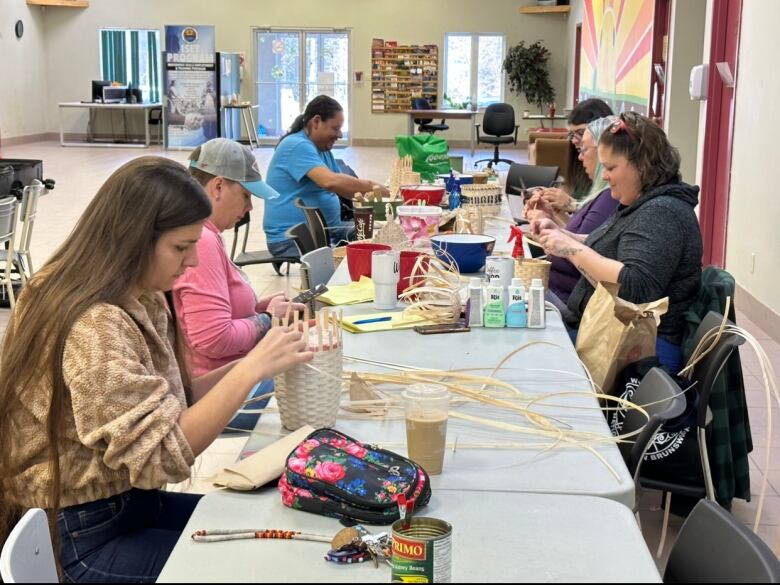 Six people sit at a long table weaving baskets.