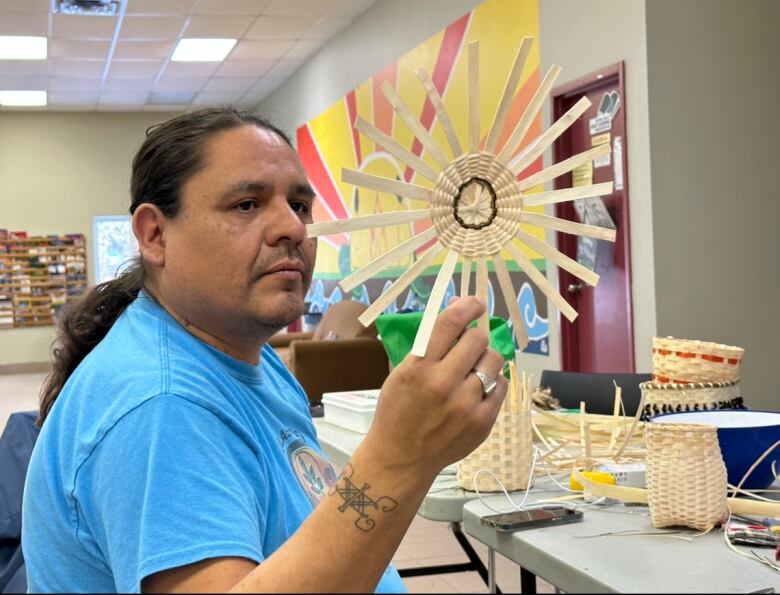 A man wearing a blue t-shirt holds up the circle-shaped bottom of an unfinished basket.