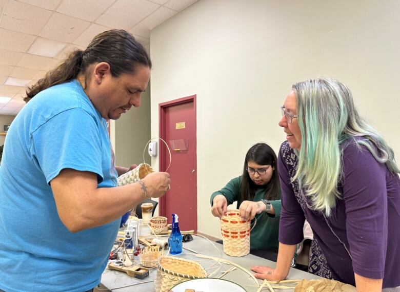 A man demonstrates to a woman how to weave a wooden strip through a basket. In the background, another man sits at a table weaving a basket.