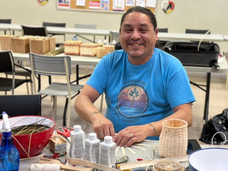 A man in a blue t-shirt smiles, his hands holding wooden strips for a small basket that sits on a table in front of him.