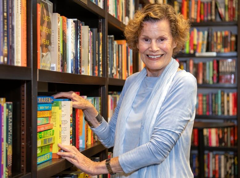 A woman wearing a pale blue outfit stands in front of a bookshelf.