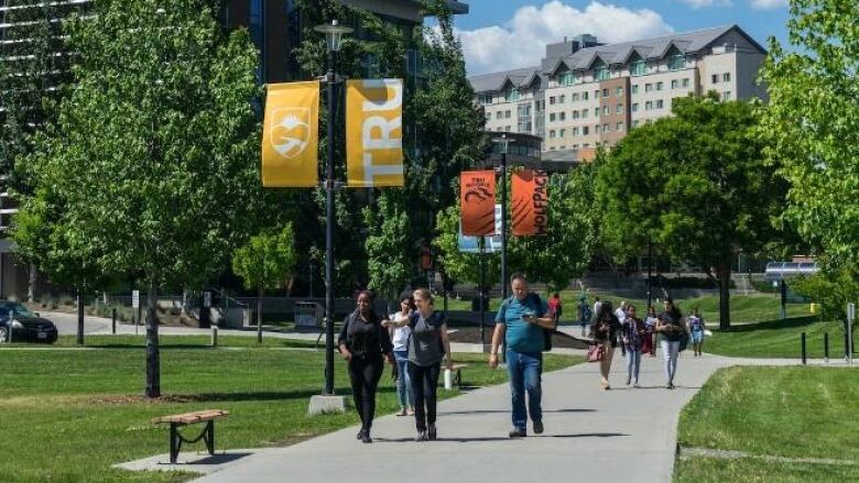 A few people are pictured walking on a pavement with grass, trees, buildings and a flagpole reading 'TRU' in the background.