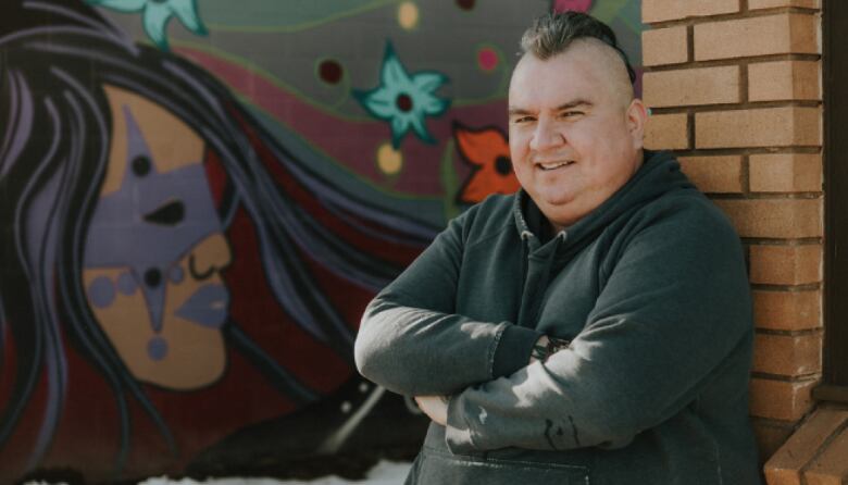 A man in black jacket crosses arms while standing near a wall.