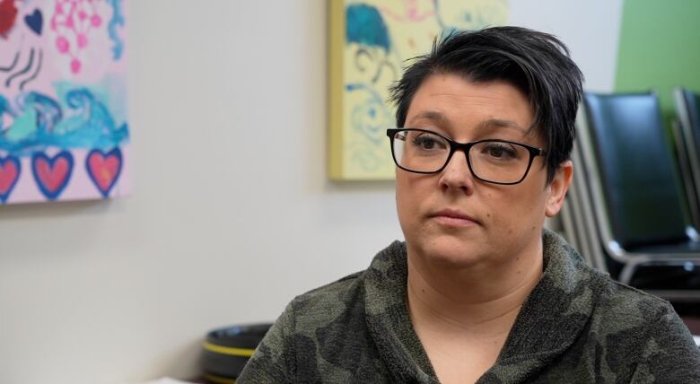 Close up of a woman with short black hear wearing glasses with colourful paintings in the background