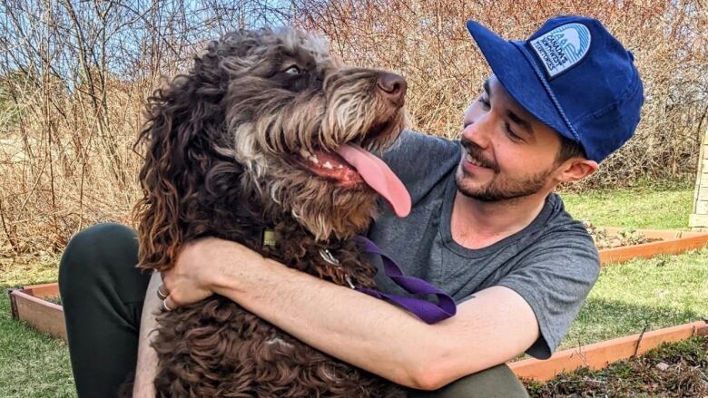 A young man wearing a baseball cap is pictured with his arms around a large dog. Both are smiling at each other.