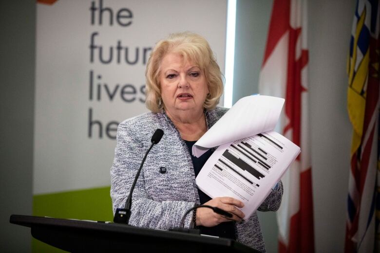 A woman with blonde shoulder-length hair standing at a podium holds open a report showing a page with blocks of blacked-out text.