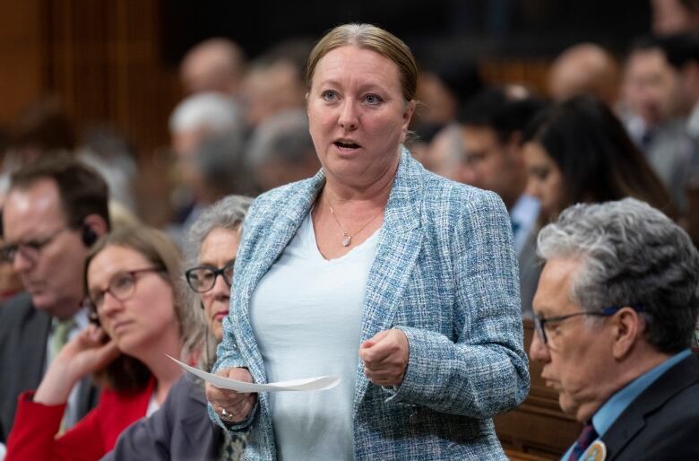 A woman wearing a blue blazer stands, holds a piece of paper and speaks to a room of people.