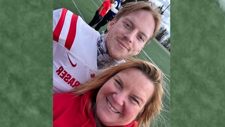 A mother and her son in a football jersey smile for the camera with a playing field in the background.