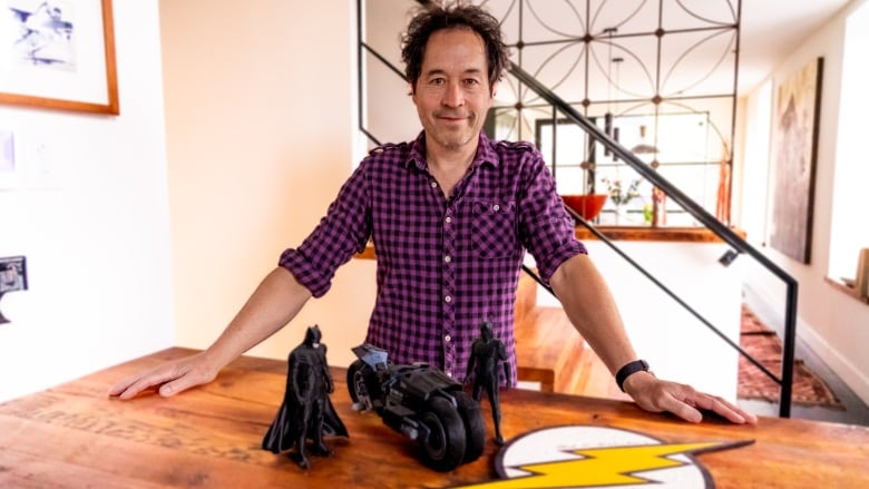 A man in a purple gingham shirt stands in a kitchen. On the table in front of him are two Batman figurines, a toy Batman motorcycle and a large emblem of The Flash. 