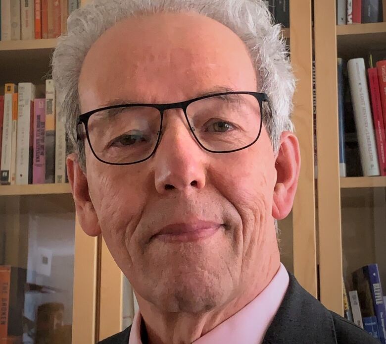 A man with short grey hair and glasses smiles in front of a bookcase.