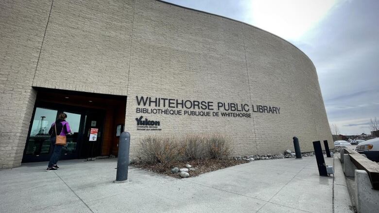 A person stands by the glass door of an otherwise grey concrete building. Thin black text on the building next to the doors says WHITEHORSE PUBLIC LIBRARY 