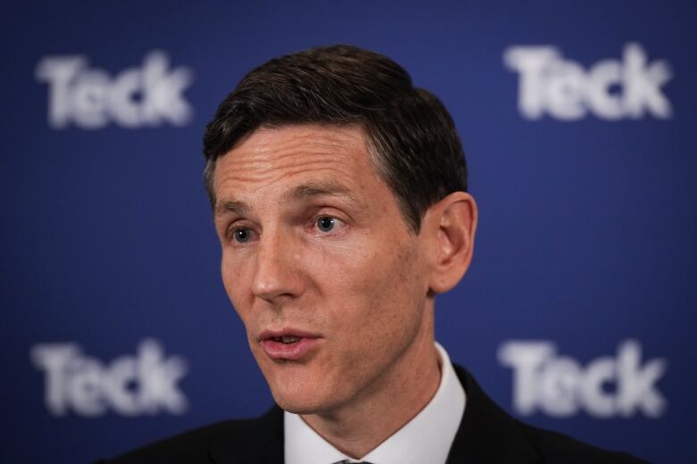 A man in a black suit stands in front of a blue board written with the words 'Teck.'