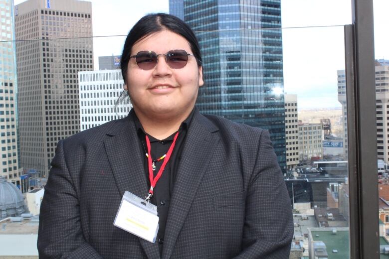 A young man with long hair and dark glasses smiles.